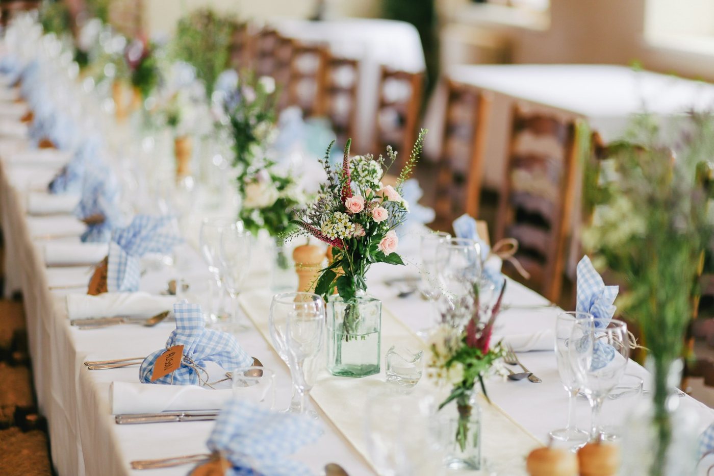A wedding reception table set with floral centerpieces featuring pink roses and mixed greenery in clear glass vases.